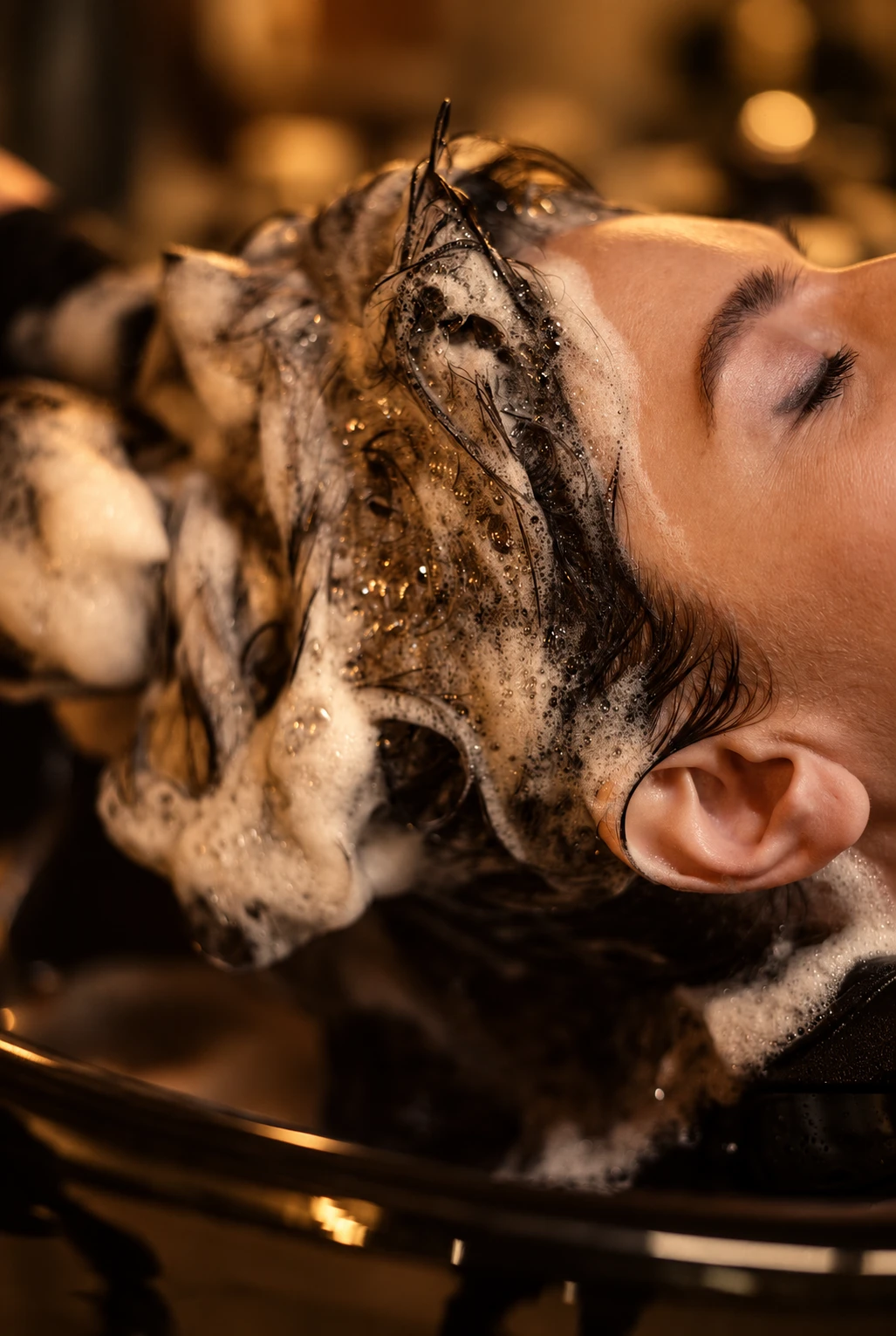 Hands lathering a client's hair in a basin during a head spa, warm golden light