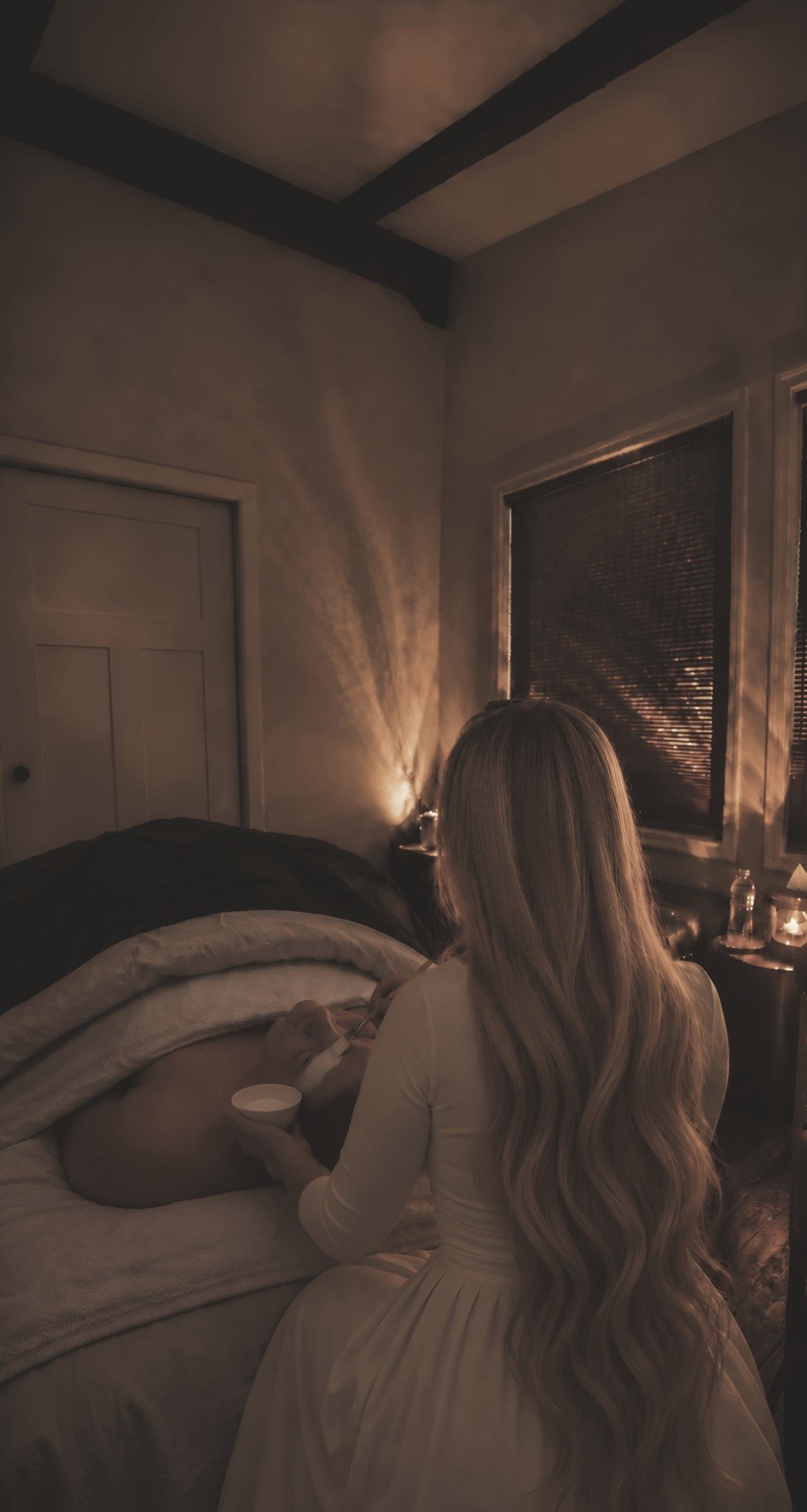 Stephanie caring for a guest in a dim, softly lit room, her long hair falling past her waist, a guest resting beneath a blanket